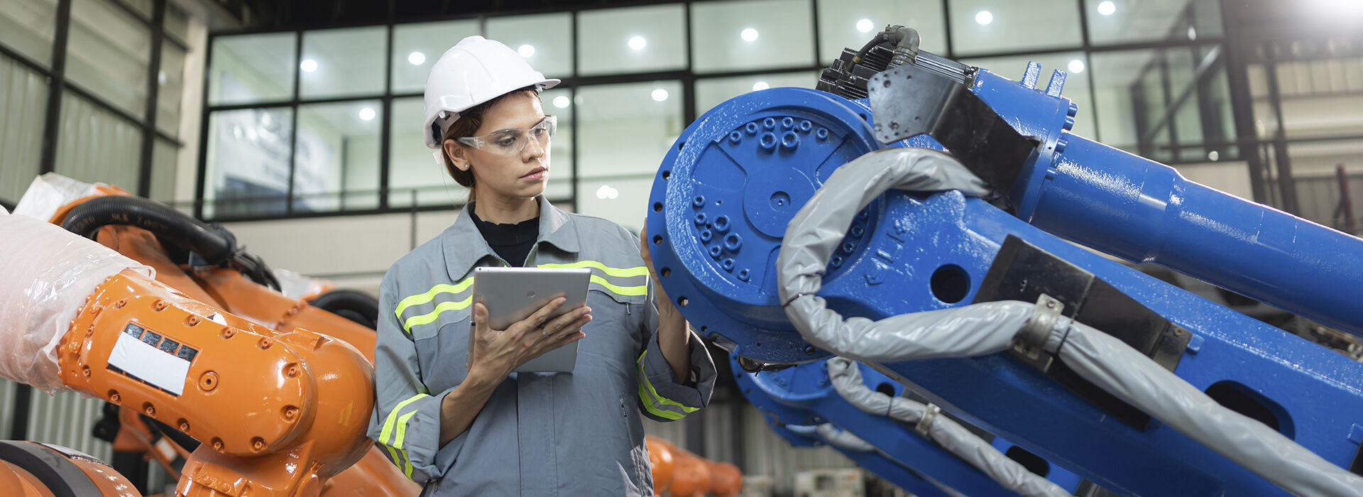 Engineer in a hard hat using a tablet to inspect an industrial robotic arm on the factory floor